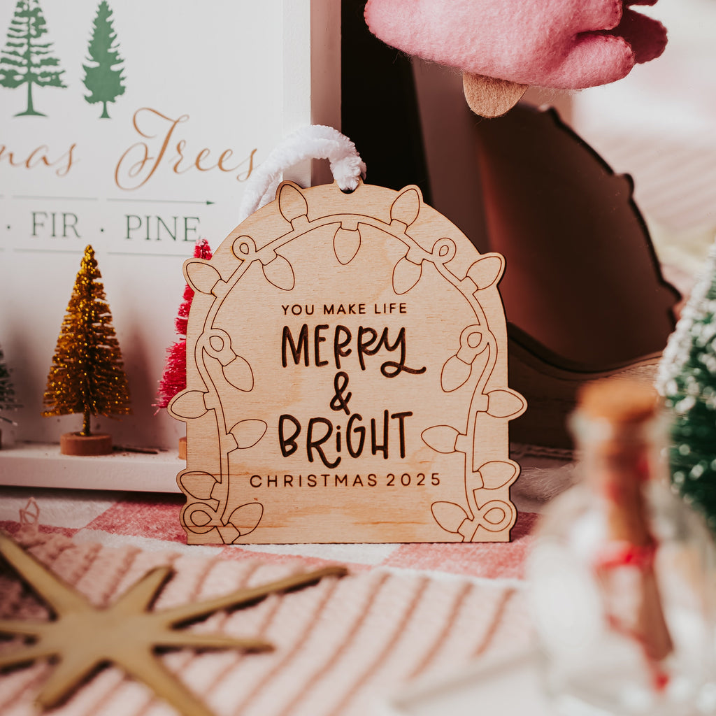A wooden Christmas ornament sign with the text 'YOU MAKE LIFE MERRY & BRIGHT CHRISTMAS 2025' displayed in a cozy, styled holiday setting with a pink felt reindeer and other rustic decorations.