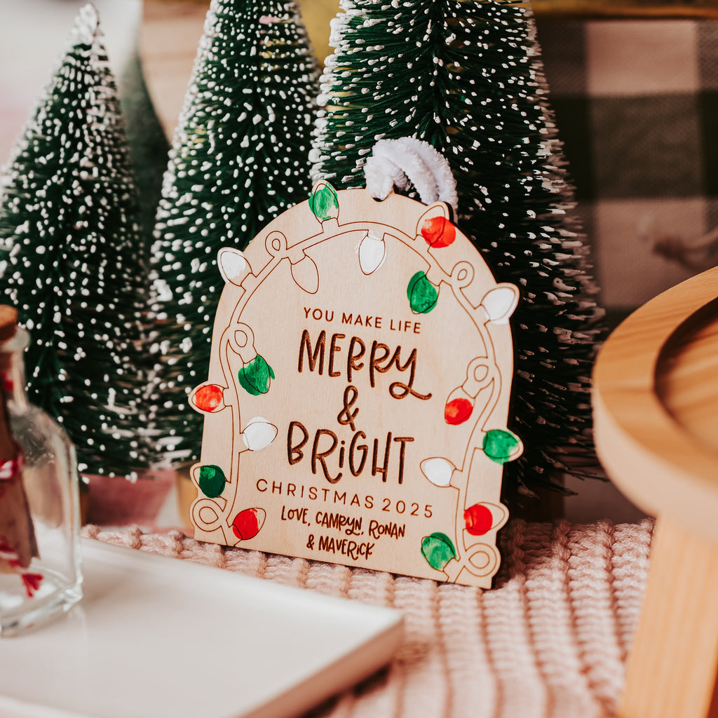 A decorative wooden ornament with a Christmas light bulb outline and the words “MERRY & BRIGHT CHRISTMAS 2025,” styled among small green bottle trees on a pink striped fabric surface.
