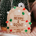 A personalized wooden Christmas ornament sign with engraved text reading 'YOU MAKE LIFE MERRY & BRIGHT CHRISTMAS 2025' and 'Love, Camryn, Ronan & Maverick,' decorated with colorful light bulbs fingerprints, shown among small green Christmas trees.