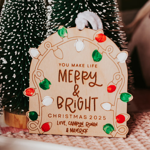A personalized wooden Christmas ornament sign with engraved text reading 'YOU MAKE LIFE MERRY & BRIGHT CHRISTMAS 2025' and 'Love, Camryn, Ronan & Maverick,' decorated with colorful light bulbs fingerprints, shown among small green Christmas trees.