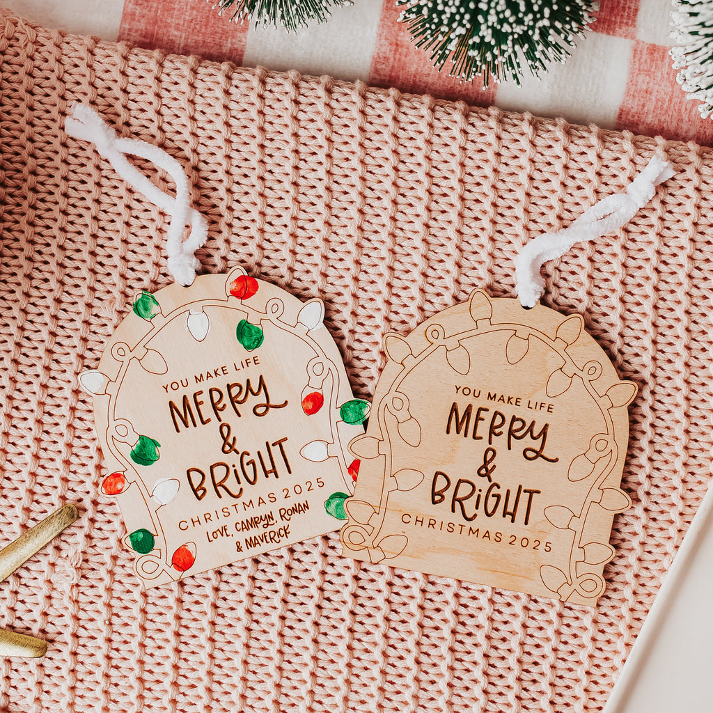 Two personalized Christmas fingerprint ornament with white hanging strings, displayed on a pink knit blanket with holiday-themed props including a starfish, a gift box, and evergreen branches.