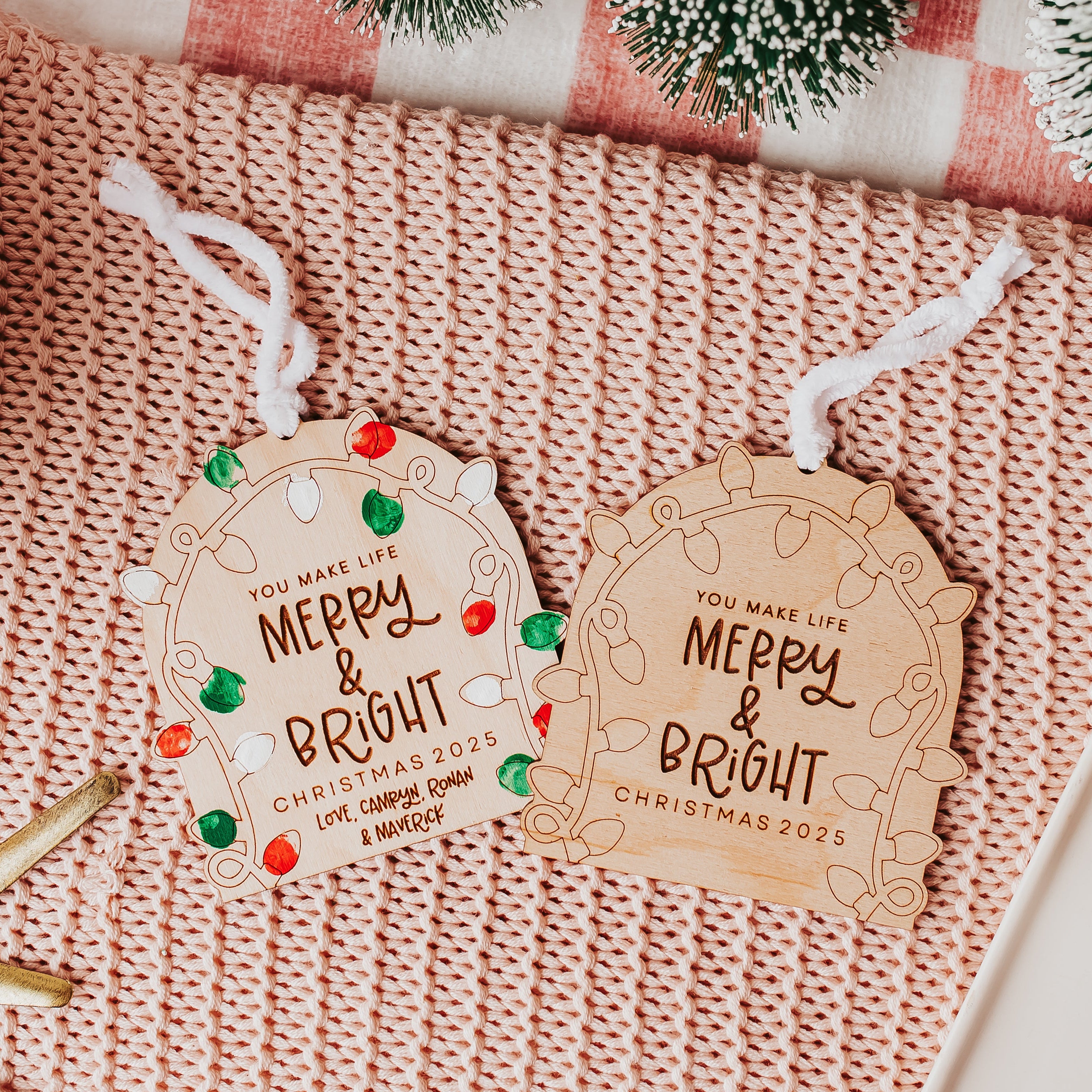 Two personalized Christmas fingerprint ornament with white hanging strings, displayed on a pink knit blanket with holiday-themed props including a starfish, a gift box, and evergreen branches.