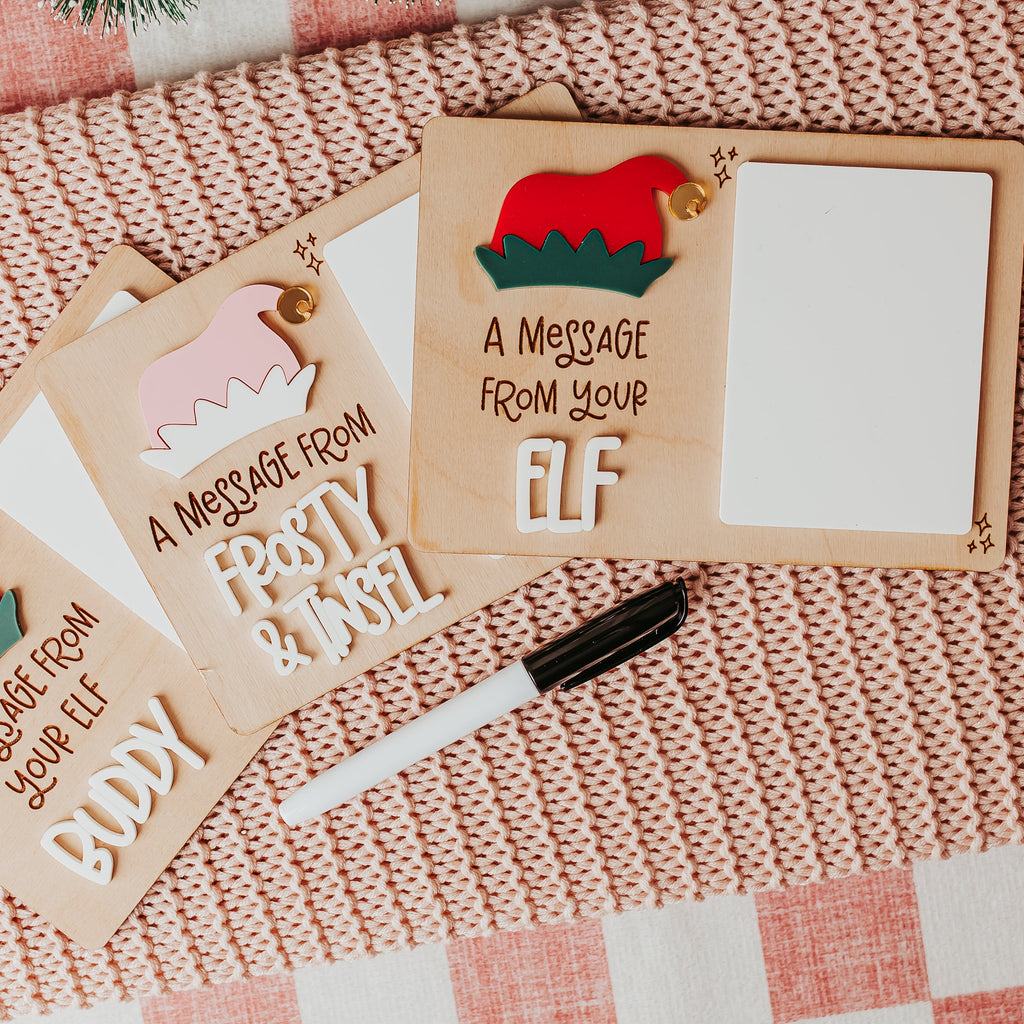 Three personalized wood and dry erase 'message from elf' signs with elf hats in pink and white and red and green, arranged on a pink knit blanket with a dry erase marker nearby.