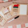 Three personalized wood and dry erase 'message from elf' signs with elf hats in pink and white and red and green, arranged on a pink knit blanket with a dry erase marker nearby.