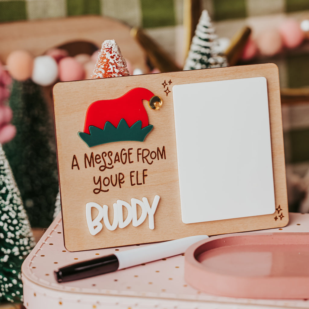 A wooden Christmas-themed message board with a red elf hat and the text 'A MESSAGE FROM YOUR ELF BUDDY' on a pink polka-dot suitcase, styled with a white marker and a pink dish in a cozy holiday setting.