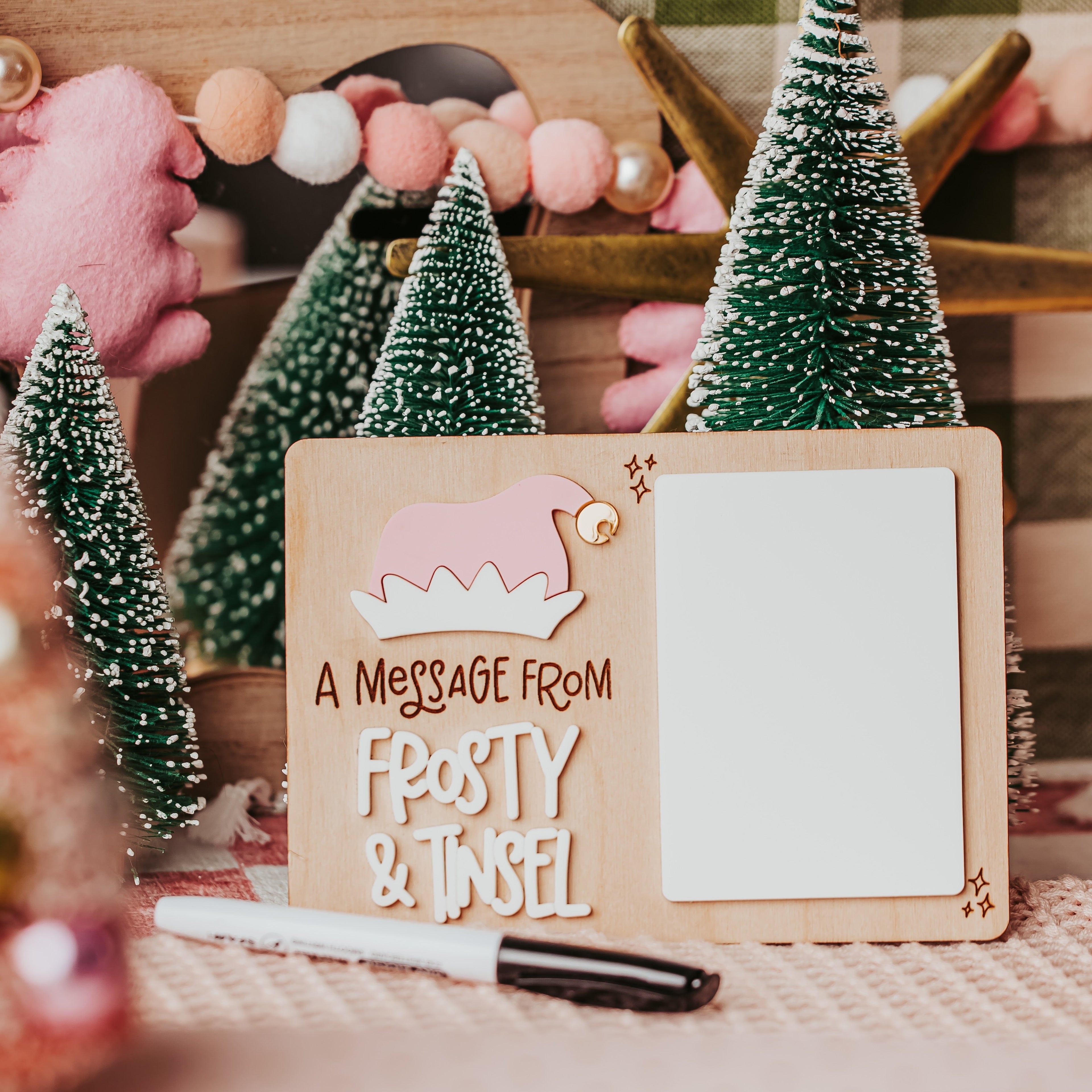 A festive wooden message card with a pink Santa hat illustration and the text 'A MESSAGE FROM FROSTY & TINSEL' on a decorated tabletop with miniature Christmas trees and a marker.
