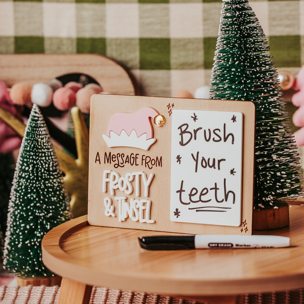 A decorative wooden sign set on a round wooden table, featuring a left sign that reads 'A MESSAGE FROM FROSTY & TINSEL' with a pink elf illustration, and a right sign that reads 'Brush Your Teeth' with small stars. A black marker lies in front.