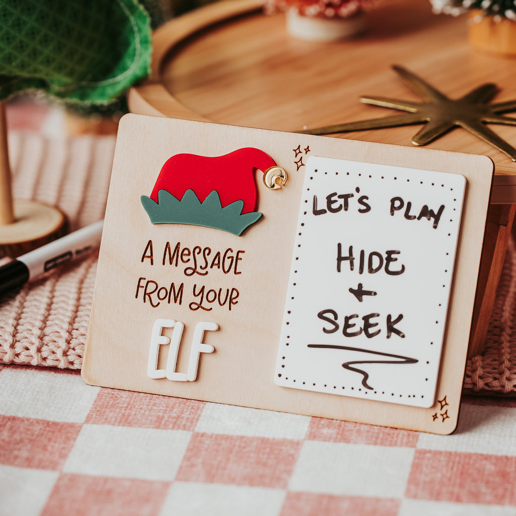 A wooden tabletop sign with a red and green elf hat illustration and the phrase 'A MESSAGE FROM YOUR ELF' on the left, next to a white card that reads 'LET'S PLAY HIDE + SEEK' on the right. The sign is styled on a pink-and-white checkered tablecloth with blurred holiday decorations in the background.