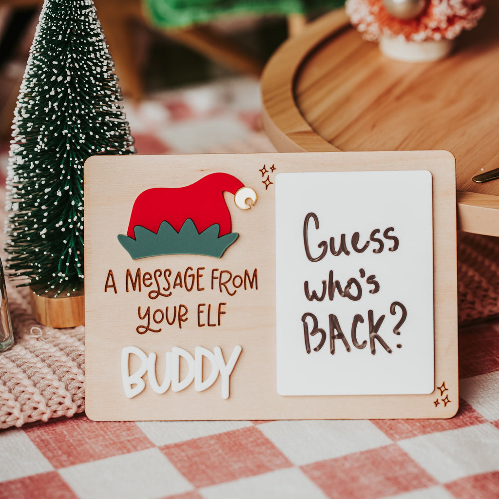 A wooden Christmas-themed sign with a red Santa hat illustration and the text 'A MESSAGE FROM YOUR ELF BUDDY' on the left, and 'Guess who's BACK?' on the right, styled on a red-and-white checkered tablecloth with miniature trees in the background.