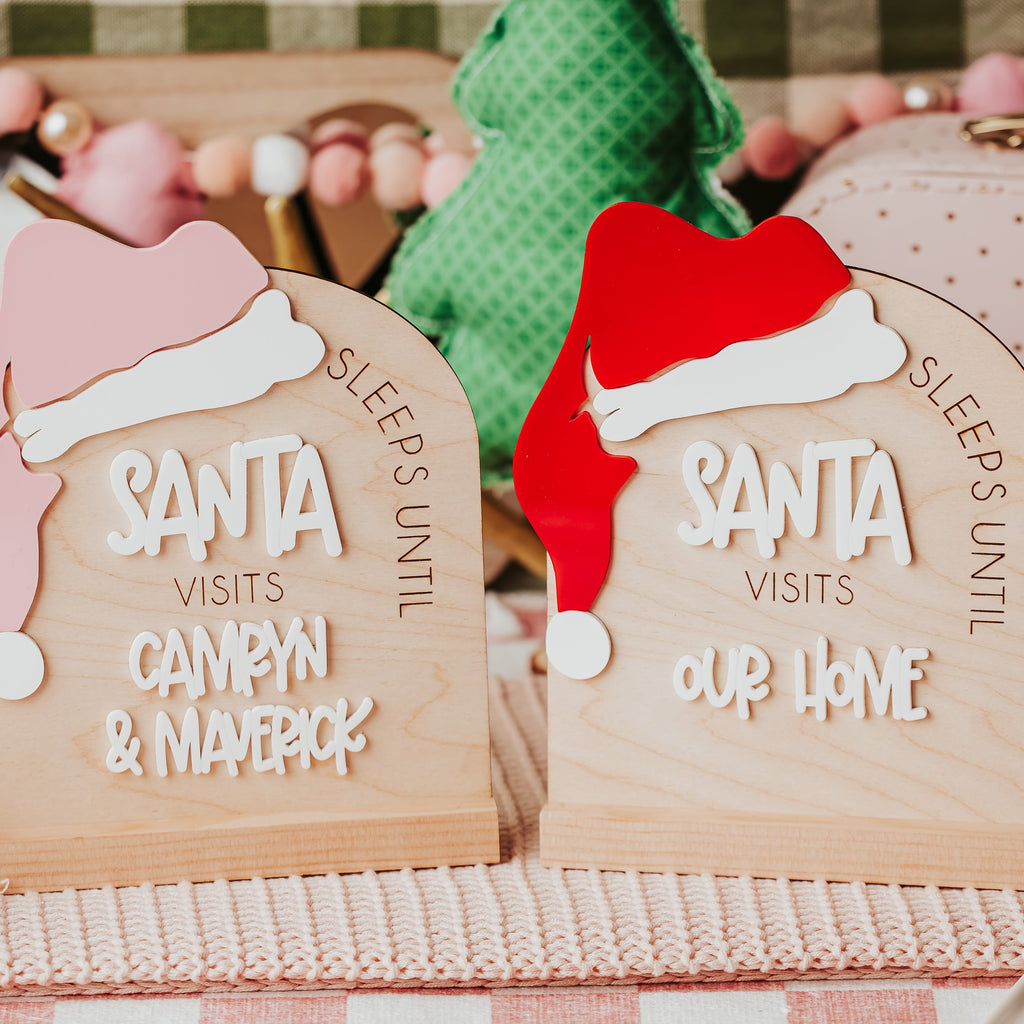 Two wooden arch Christmas sign plaques with Santa hats, one reading 'Sleeps until Santa visits Camryn & Maverick' and the other 'Santa visits our home', styled on a pink knit surface with holiday decor in the background.