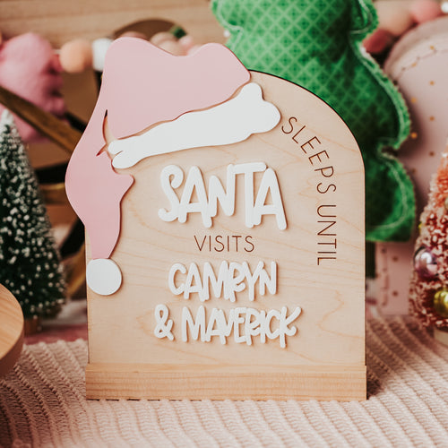A wooden sign shaped like an arch with Santa's pink hat sticking over the top , reading 'SLEEPS UNTIL SANTA VISITS CAMRYN & MAVERICK' styled on a pink checkered surface with holiday decorations in the background.
