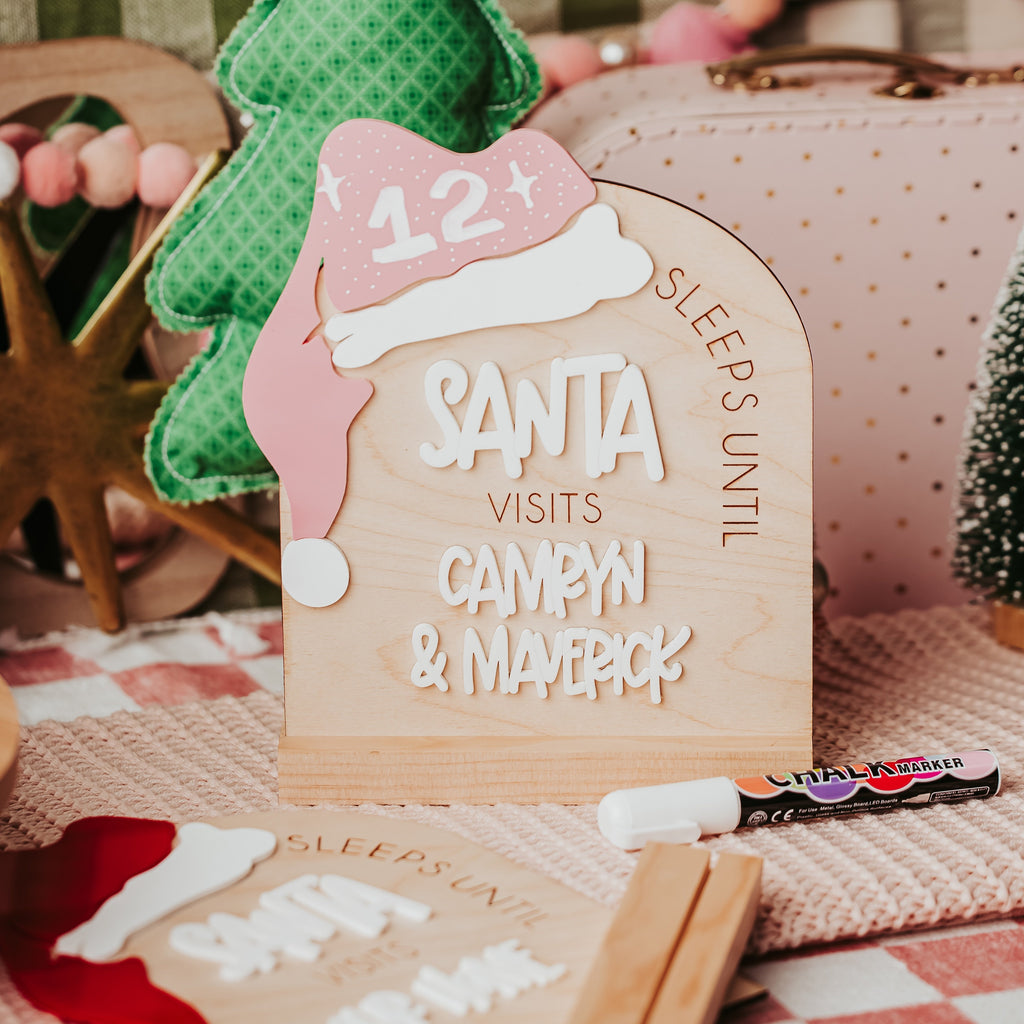 A wooden arch Christmas with a Santa hat over the top that reads 'SLEEPS UNTIL SANTA VISITS CAMRYN & MAVERICK'. Styled with a green felt Christmas tree, pink polka-dot bag, and holiday decorations in the background.