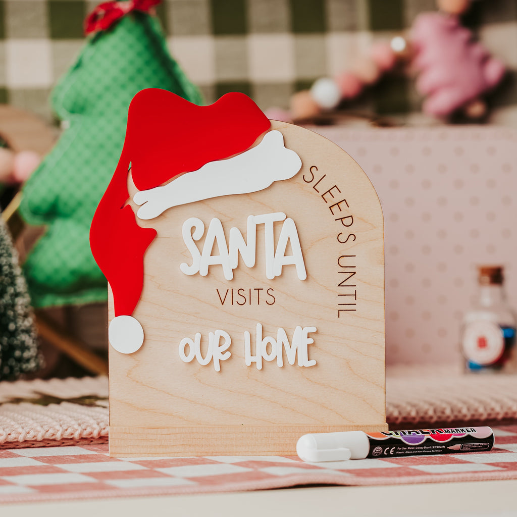 A wooden arch Christmas sign a red Santa hat, reading 'SLEEPS UNTIL SANTA VISITS OUR HOME' displayed on a table with small tree decorations and a marker in a cozy indoor setting.