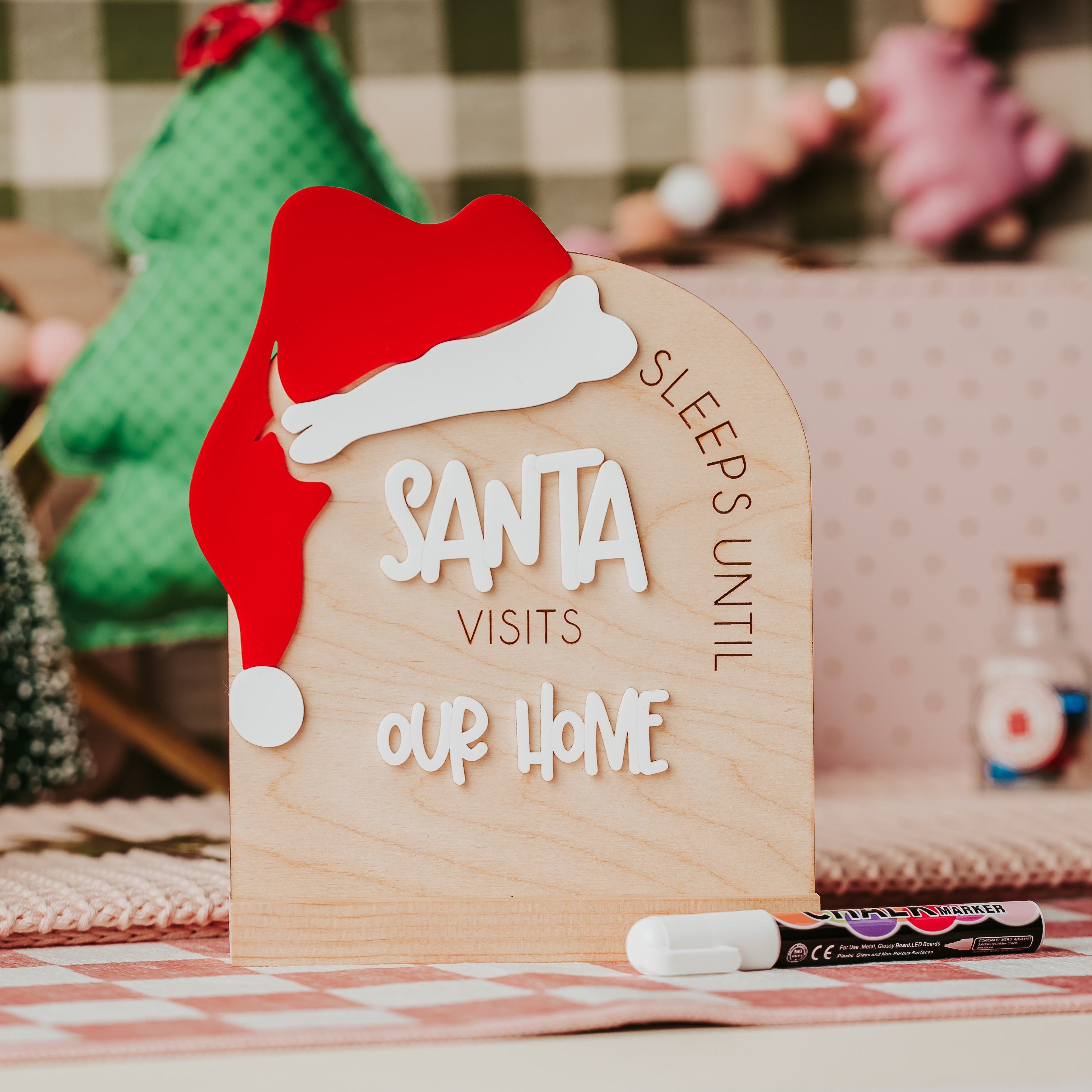 A wooden arch Christmas sign a red Santa hat, reading 'SLEEPS UNTIL SANTA VISITS OUR HOME' displayed on a table with small tree decorations and a marker in a cozy indoor setting.