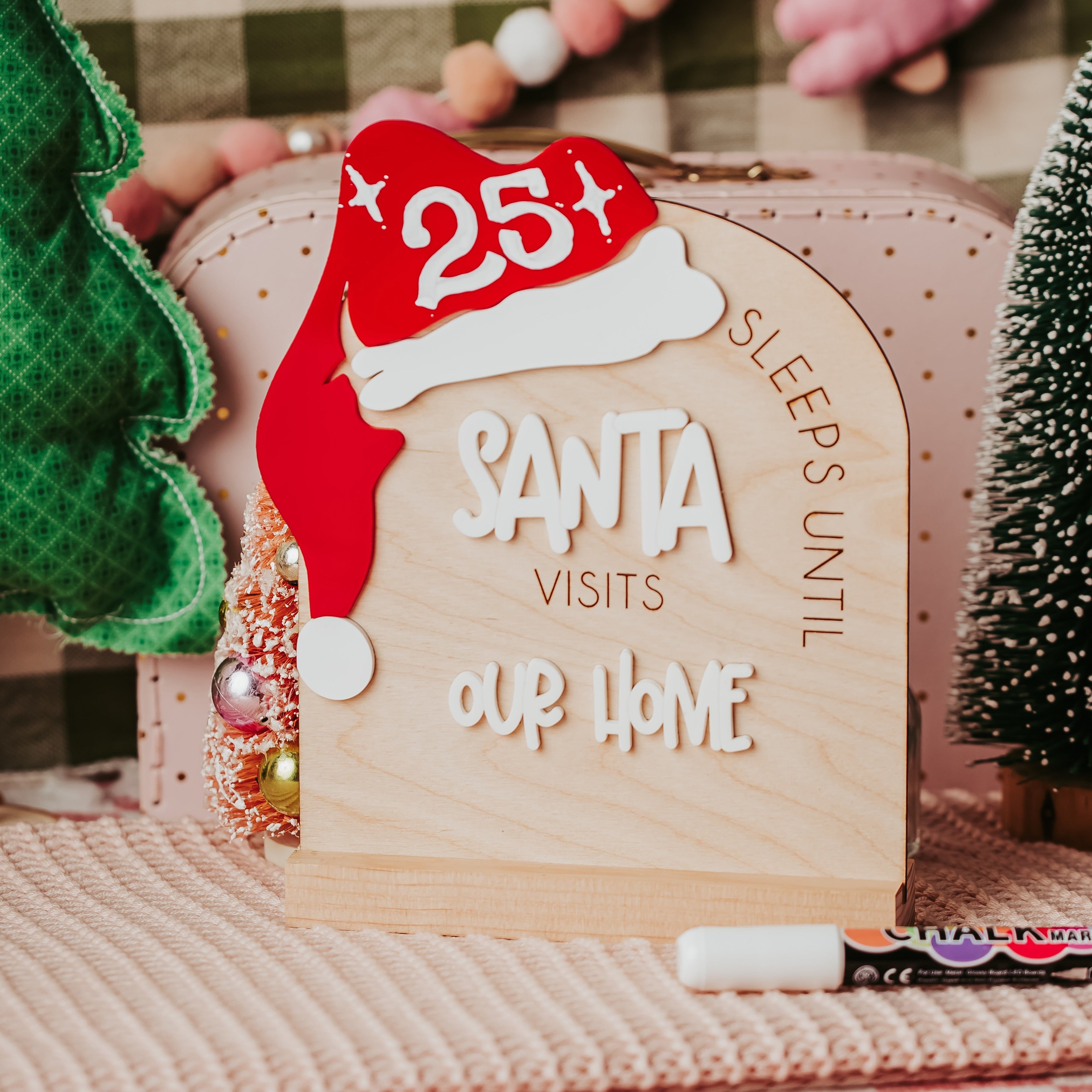 A wooden arch Christmas sign with a red Santa hat and white lettering that reads 'SLEEPS UNTIL SANTA VISITS OUR HOME'. The sign is styled on a pink checkered blanket with a pink polka-dot suitcase, a green plush tree, and a pink beaded necklace in the background.