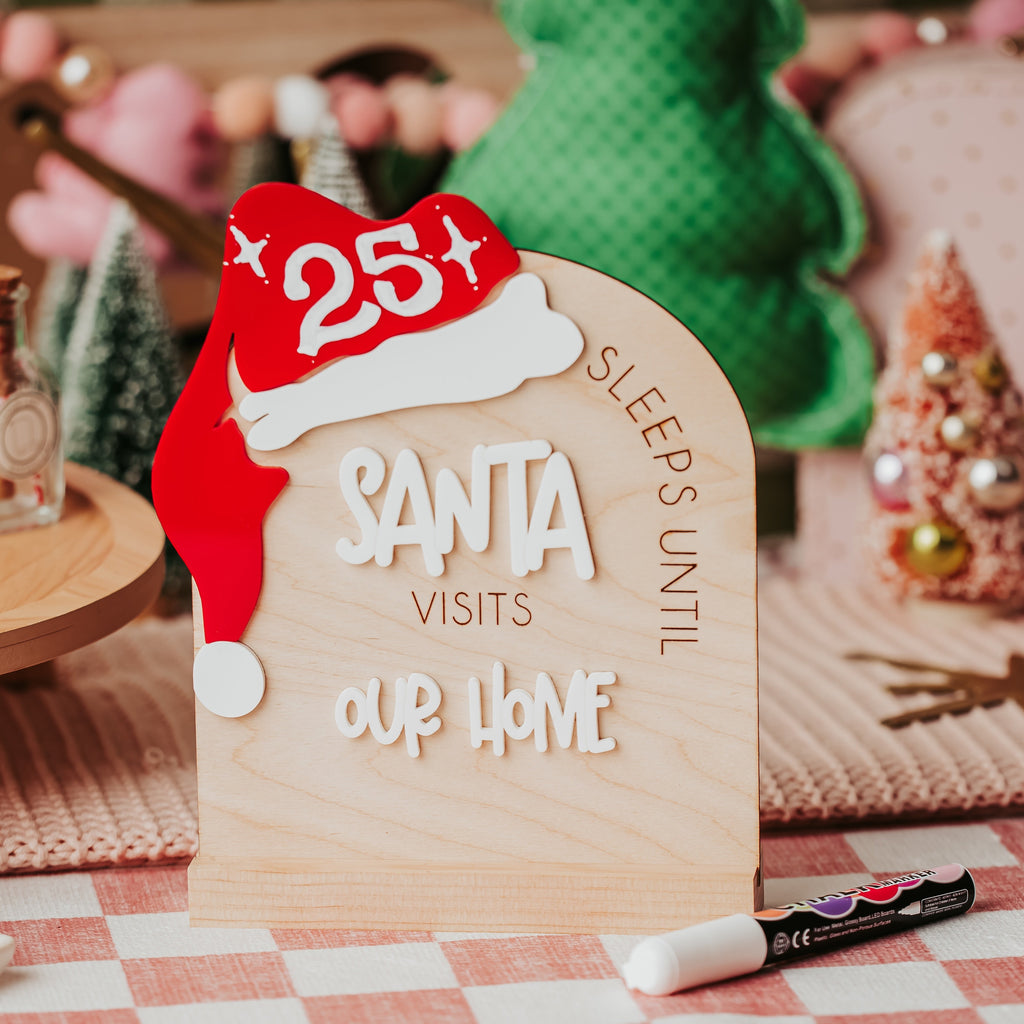 A wooden arch Christmas sign with a red Santa hat and the words 'SLEEPS UNTIL SANTA VISITS OUR HOME', styled on a checkered tablecloth with holiday decor in the background.