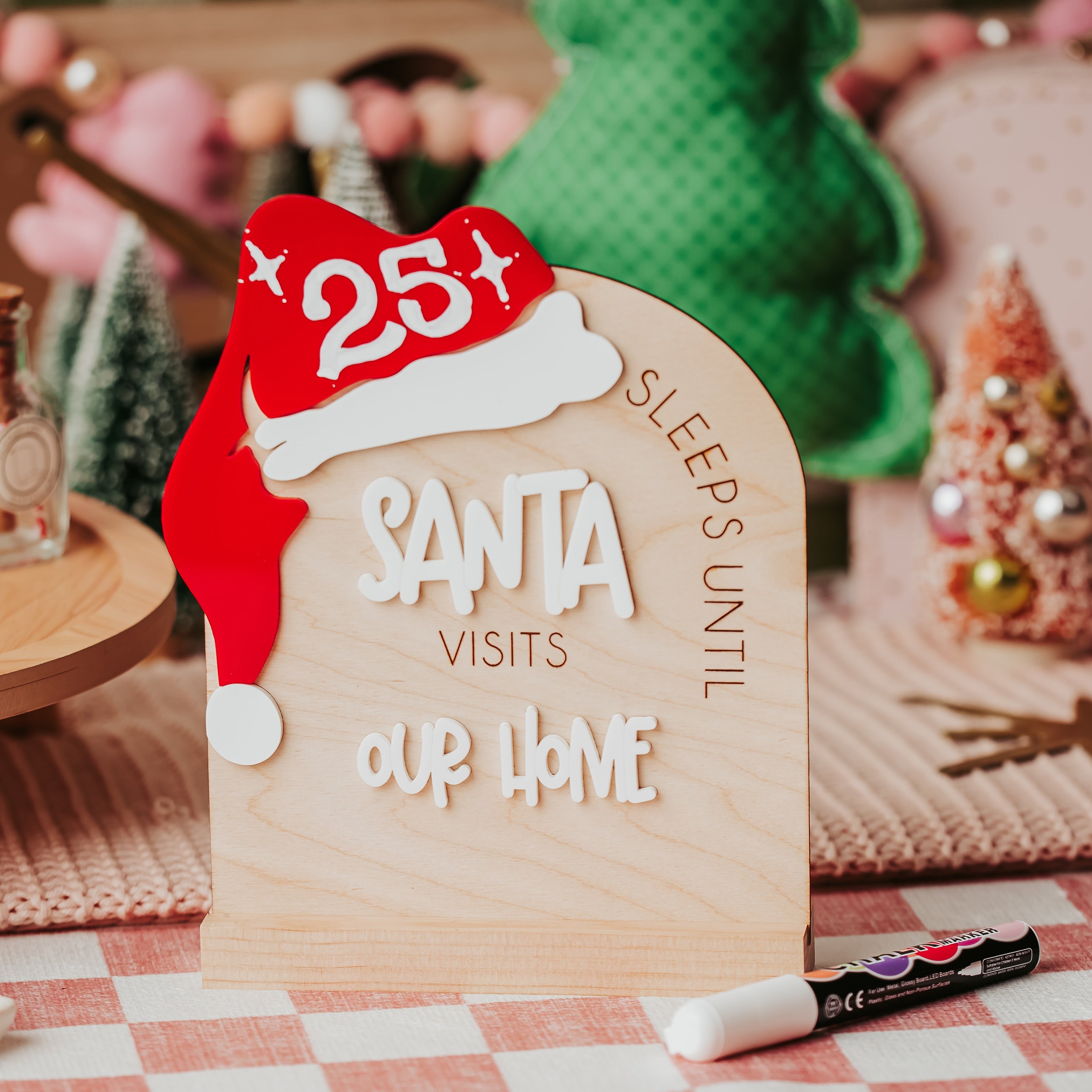 A wooden arch Christmas sign with a red Santa hat and the words 'SLEEPS UNTIL SANTA VISITS OUR HOME', styled on a checkered tablecloth with holiday decor in the background.
