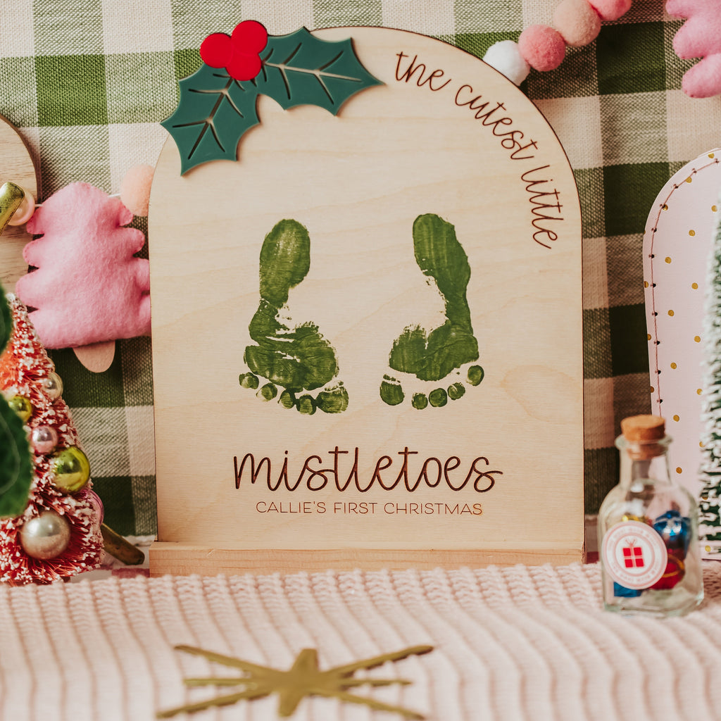 A personalized wooden keepsake sign with wooden stand featuring green baby footprints and the text 'the cutest little mistletoes CALLIE'S FIRST CHRISTMAS' surrounded by festive Christmas decorations, including a green felt tree, a small pink plush hand, and miniature trees.