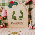 A personalized wooden keepsake sign with wooden stand featuring green baby footprints and the text 'the cutest little mistletoes CALLIE'S FIRST CHRISTMAS' surrounded by festive Christmas decorations, including a green felt tree, a small pink plush hand, and miniature trees.