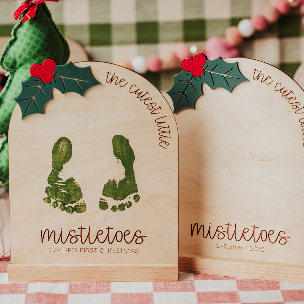 Two personalized wooden baby footprint signs for a child's first Christmas, decorated with green holly leaves and red berries , standing on a checkered tablecloth with miniature Christmas trees in the background.
