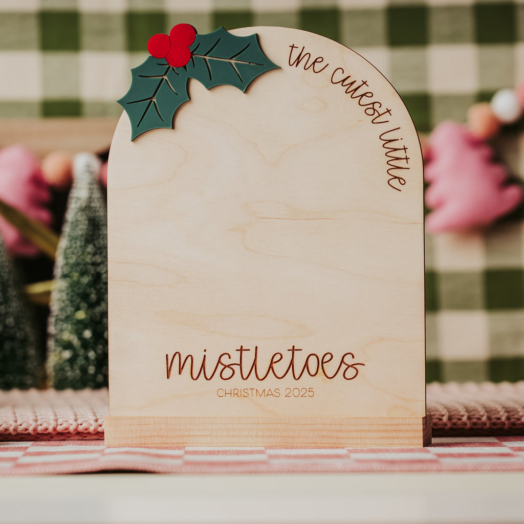 A wooden arch shaped Christmas sign with a green holly leaf and red berries at the top, and the words 'the cutest little mistletoes' and 'mistletoes CHRISTMAS 2025' engraved on it, displayed on a decorated table with festive background elements. Sign is designed to place painted baby footprints on it for a Christmas keepsake.
