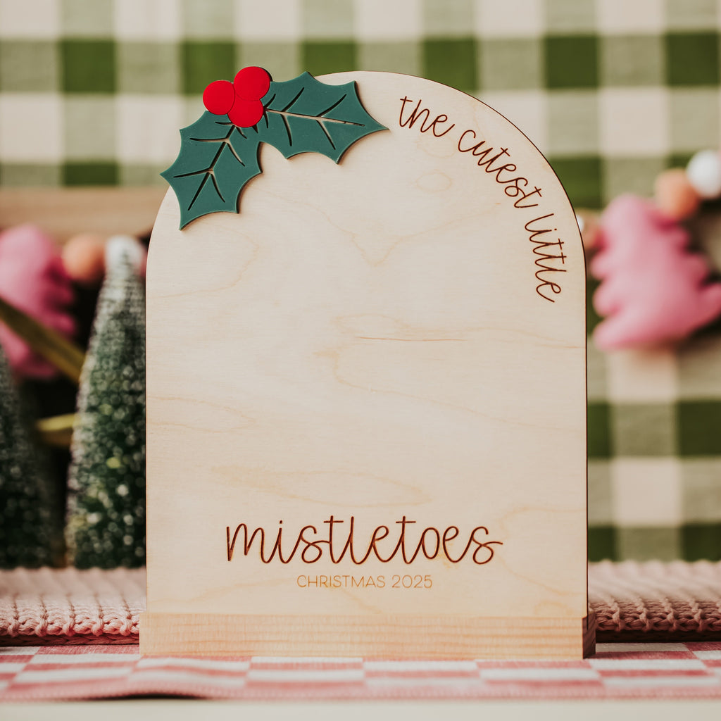 A personalized wooden Christmas table sign with an arched top, featuring a green holly leaf and red heart decoration at the top left. The sign is engraved with 'the cutest little mistletoes' on the upper right and 'mistletoes CHRISTMAS 2025' on the lower front.