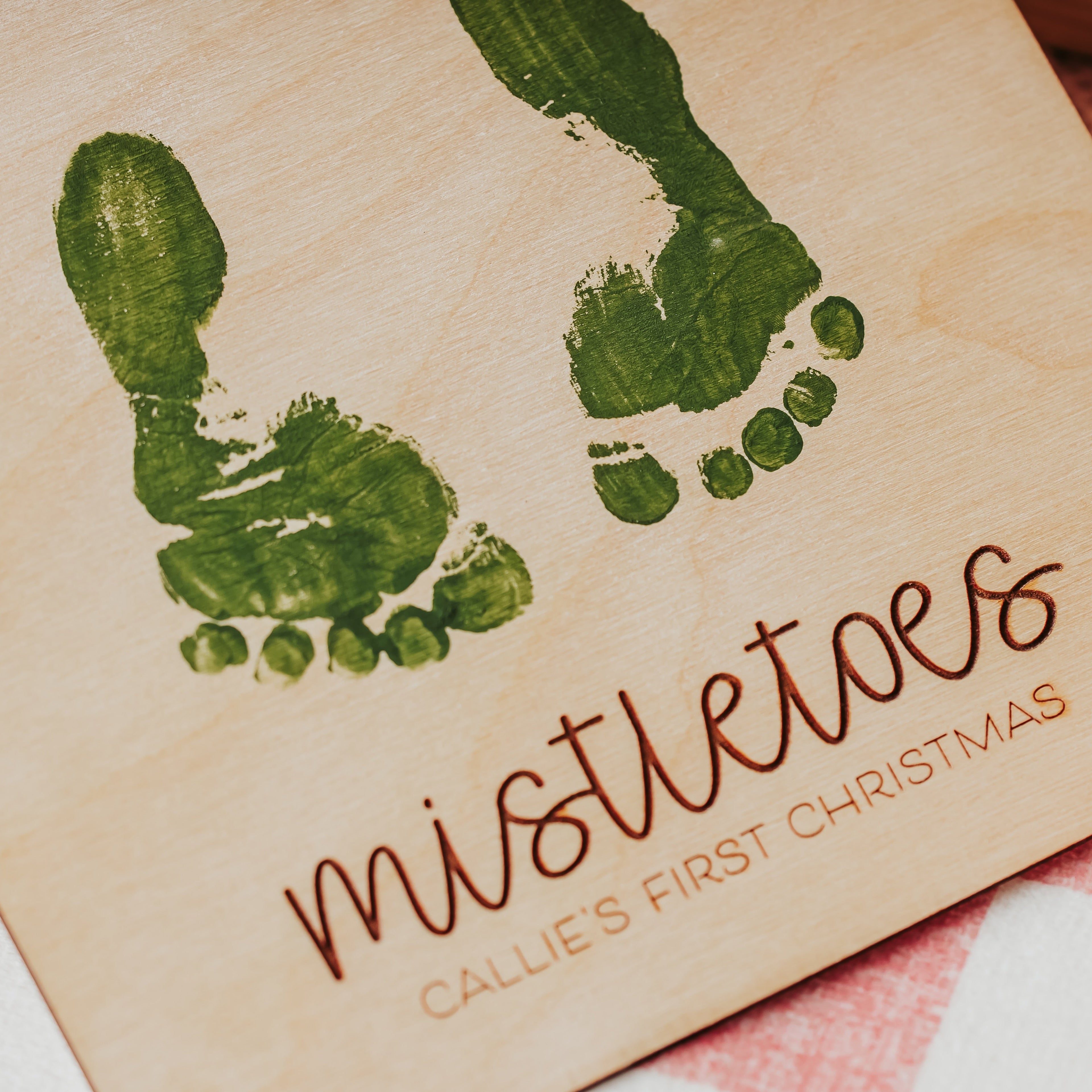 A close-up of a personalized wooden plaque featuring green painted baby footprints and the words 'mistletoes CALLIE'S FIRST CHRISTMAS' in brown script, resting on a pink-and-white checkered fabric surface.