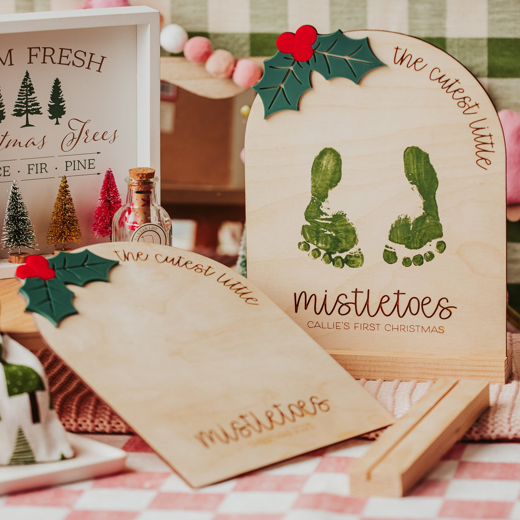 Personalized wooden baby footprint keepsake sign. on a red-and-white checkered blanket, featuring green footprint cutouts and the words 'we cut out little mistletoes' with holly accents. 