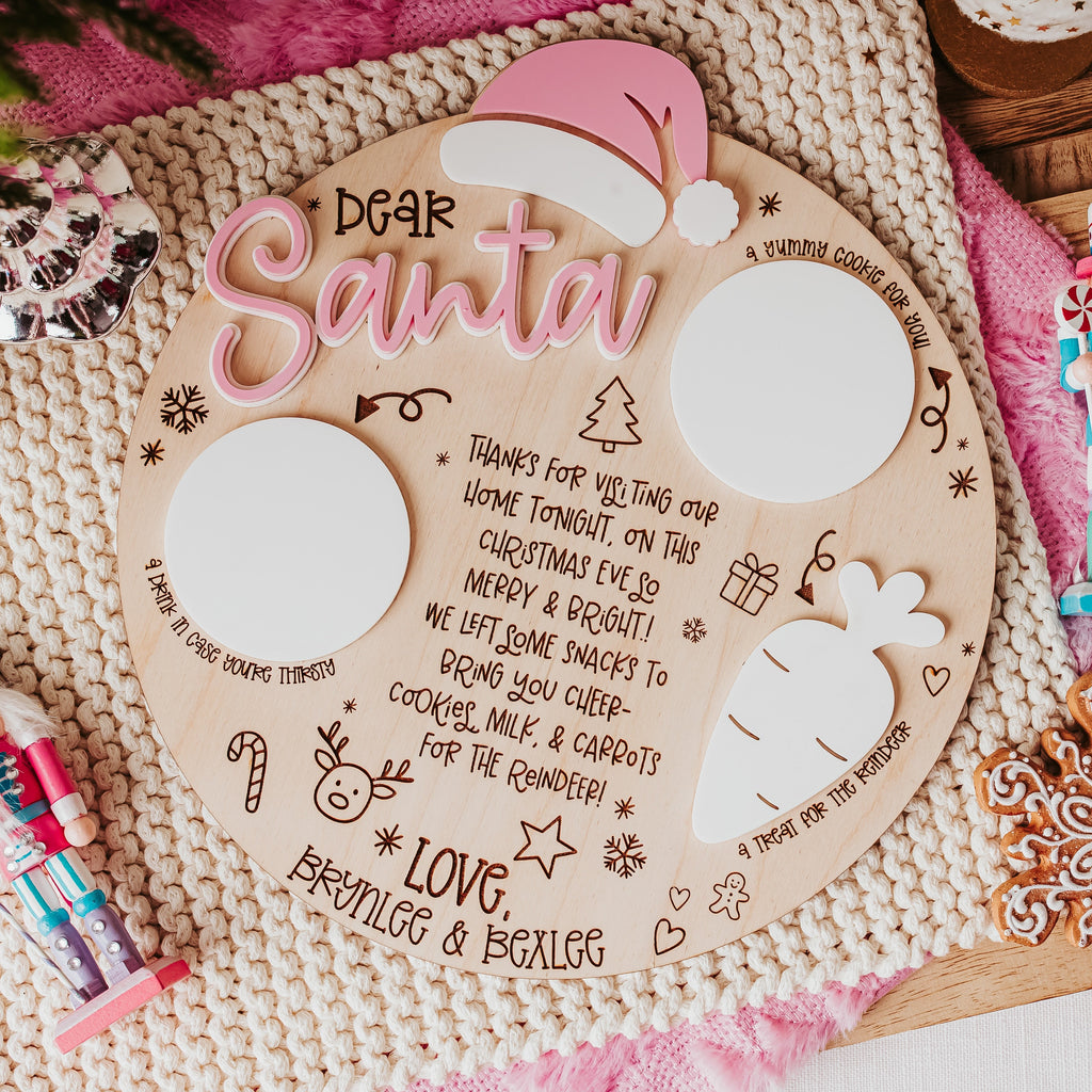 A festive flat-lay of a personalized wooden Christmas tray with 'Dear Santa' written in pink script. The has two blank white circles for milk & cookies for santa and a white carrot to place carrots for the reindeers and, decorated with hand-drawn illustrations of a carrot, candy cane, and Christmas tree.