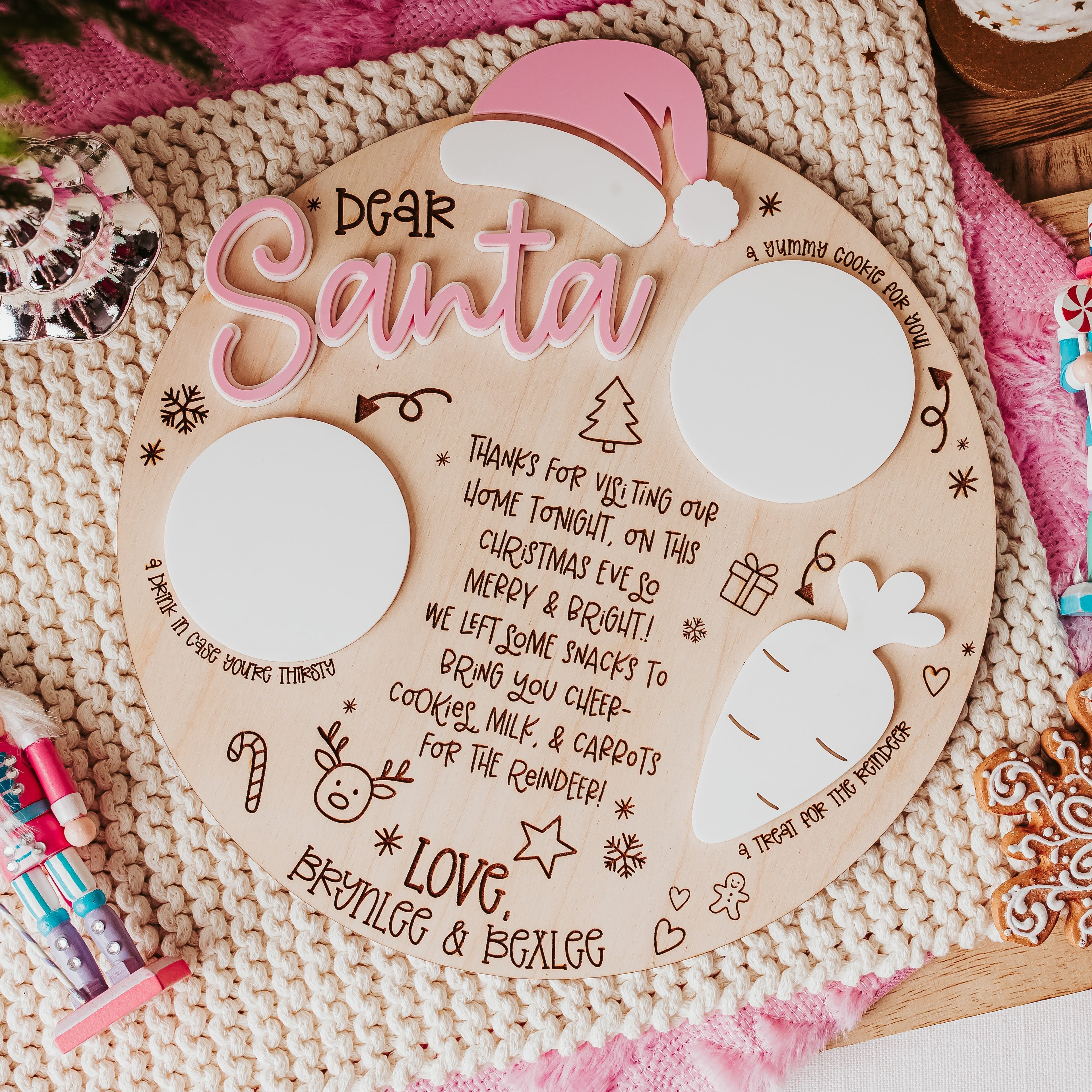 A festive flat-lay of a personalized wooden Christmas tray with 'Dear Santa' written in pink script. The has two blank white circles for milk & cookies for santa and a white carrot to place carrots for the reindeers and, decorated with hand-drawn illustrations of a carrot, candy cane, and Christmas tree.