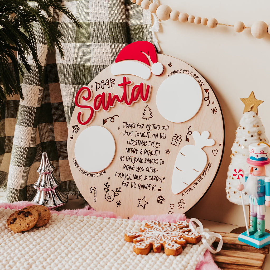 A round wooden Christmas tray with a red Santa hat,  in a cozy holiday setting with plaid curtains, bead garland, cookies, and small decorative ornaments.