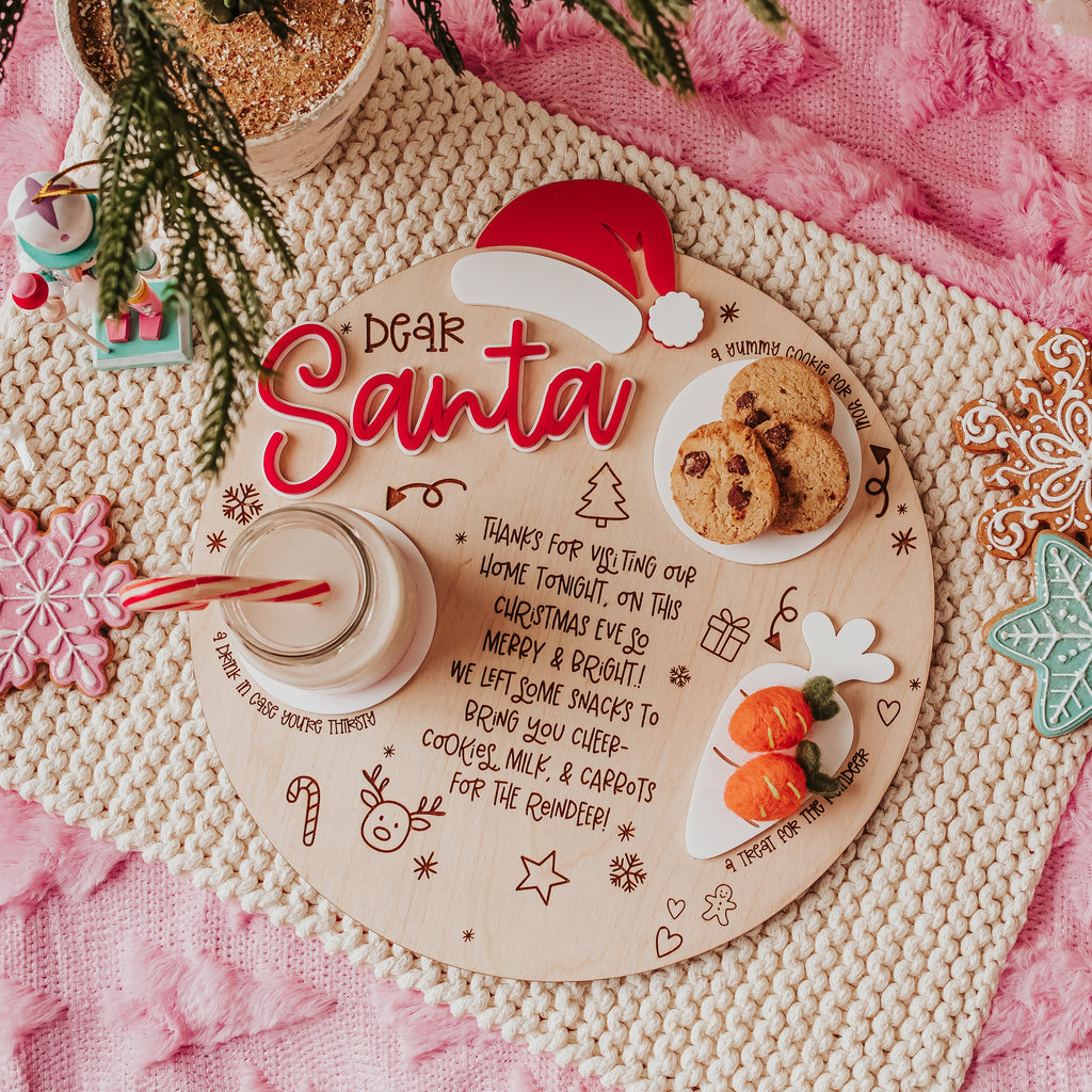 A festive holiday flat-lay featuring a personalized wooden Santa tray with 'Dear Santa' lettering, decorated with cookies, a red Santa hat, and candy canes, styled on a pink blanket with a knitted placemat, pine branches, and a plaid pillow.