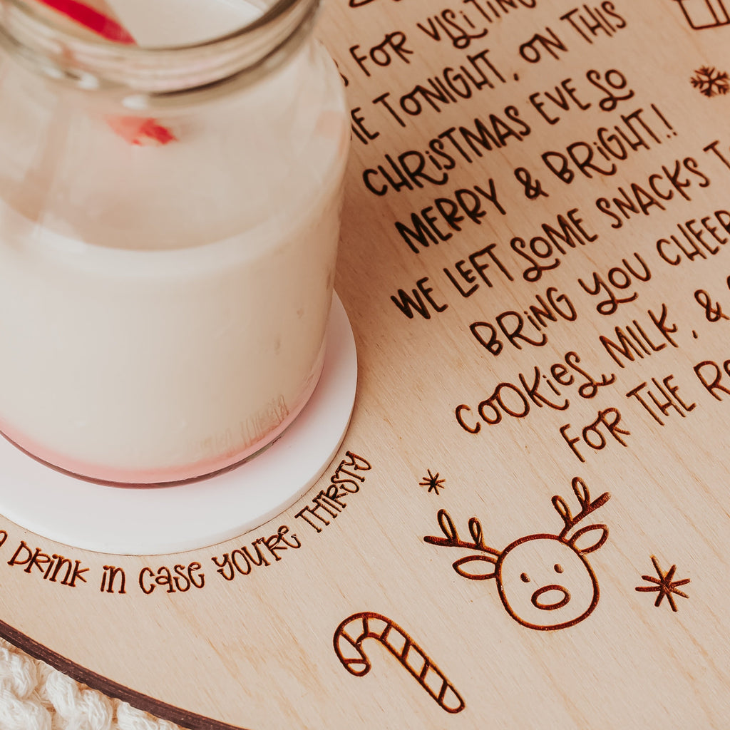 A close-up of a wooden Christmas-themed tray or engraved holiday text and illustrations, holding a glass of milk with a red-and-white striped straw, with cookies on a plate nearby.