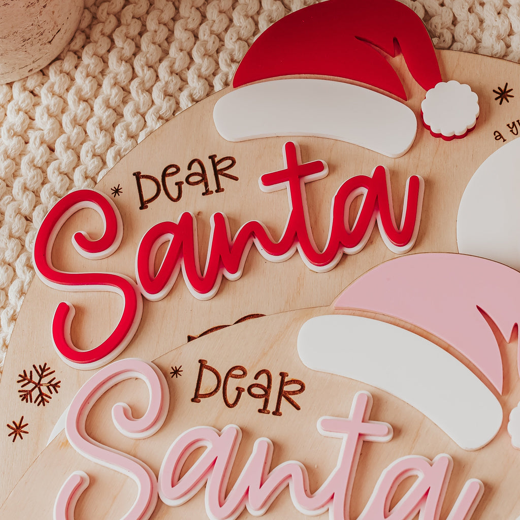 A close-up of layered wooden Christmas tray reading 'DEAR Santa' in red and pink, decorated with a Santa hat silhouette, snowflakes, and small holiday icons on a light wood background.