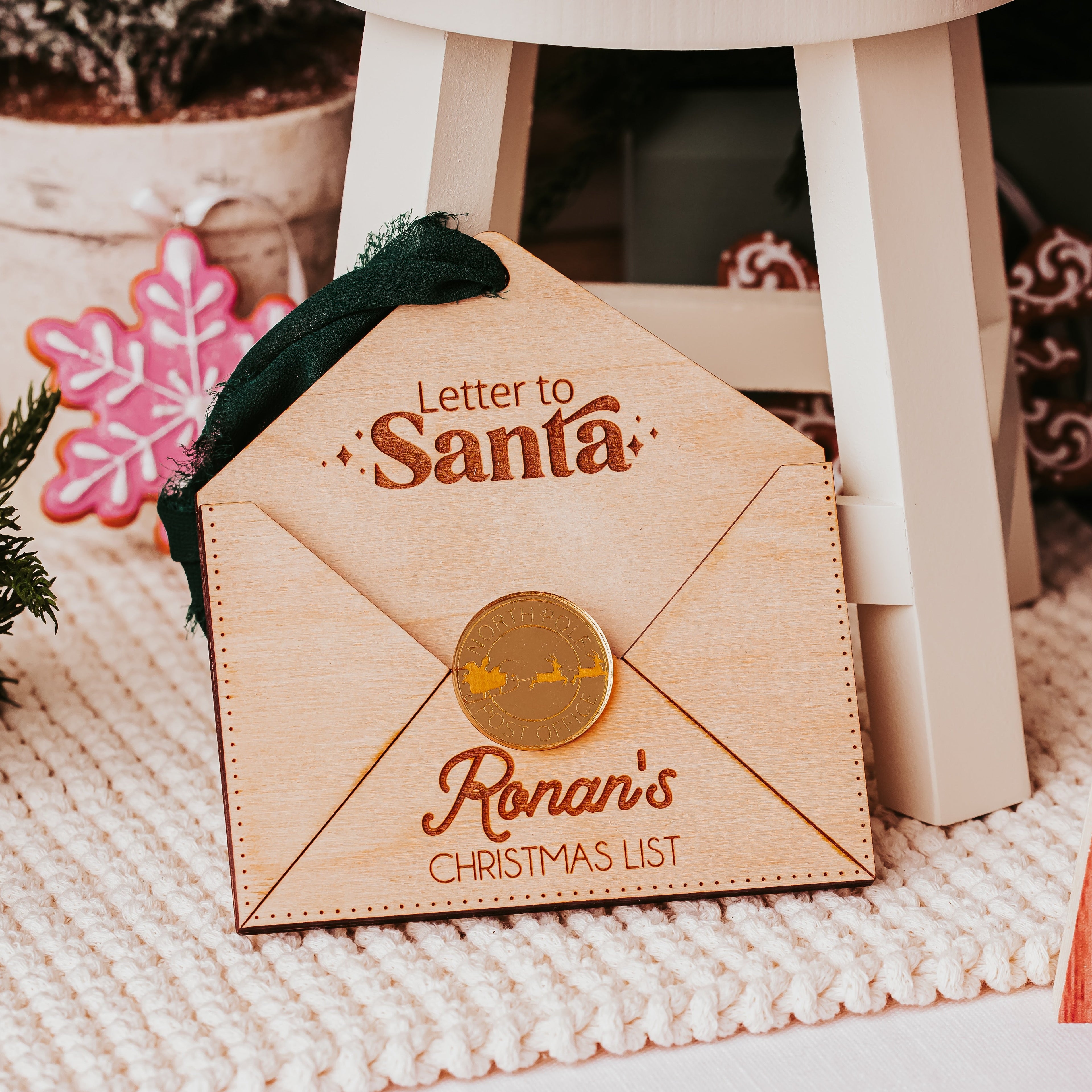 A wooden Christmas envelope with a printed letter to Santa and the name 'Ronan's Christmas List' on the front, styled on a white knit blanket with holiday decor in the background.