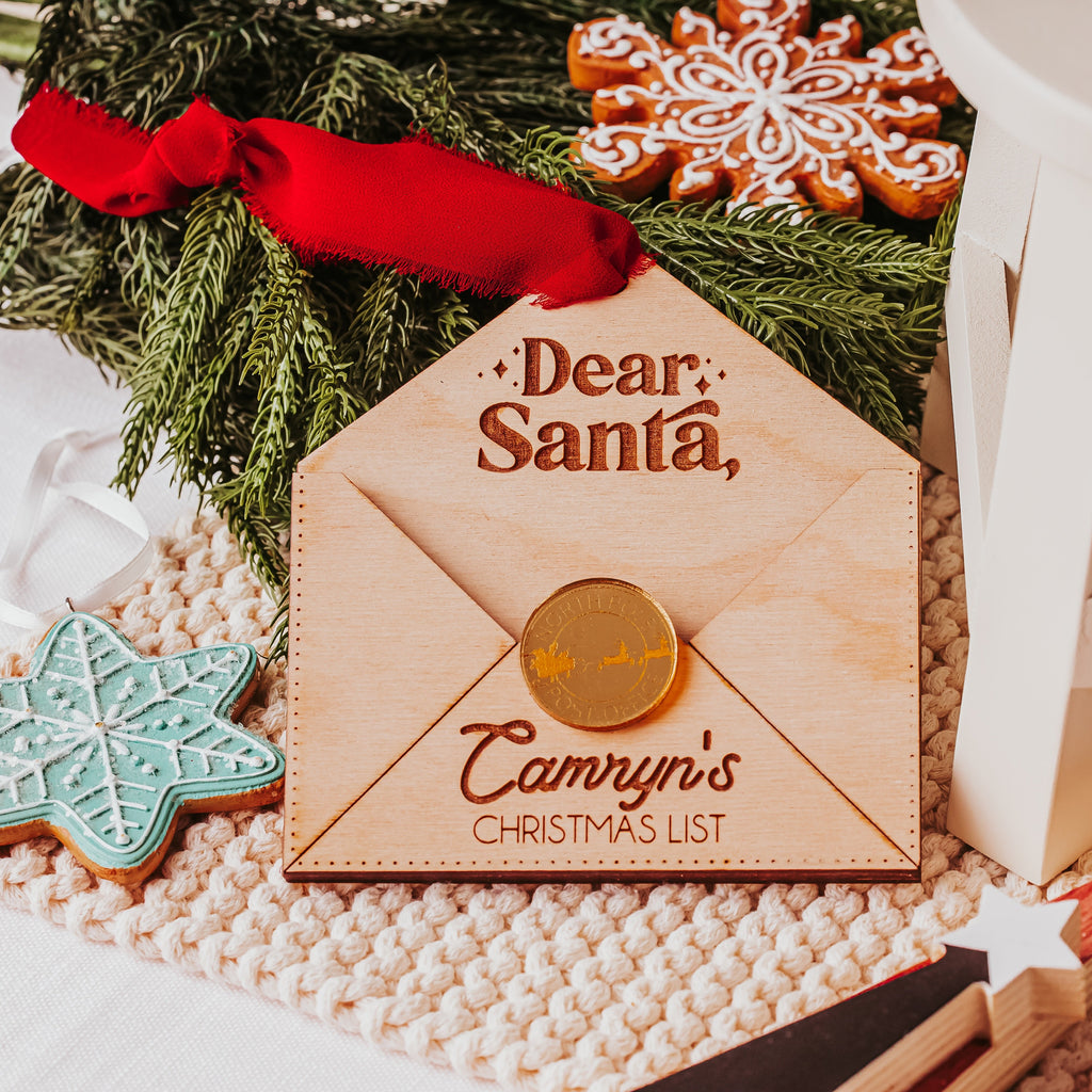 A personalized wooden Christmas list envelope with a red ribbon and acrylic gold wax seal, styled on a knit blanket with holiday cookies and greenery.