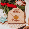 A personalized wooden Christmas list envelope with a red ribbon and acrylic gold wax seal, styled on a knit blanket with holiday cookies and greenery.