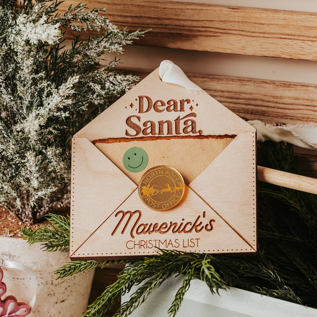 A personalized wooden Christmas envelope ornament with green pine branches, a wooden bead garland, and a white pot in a cozy indoor setting.