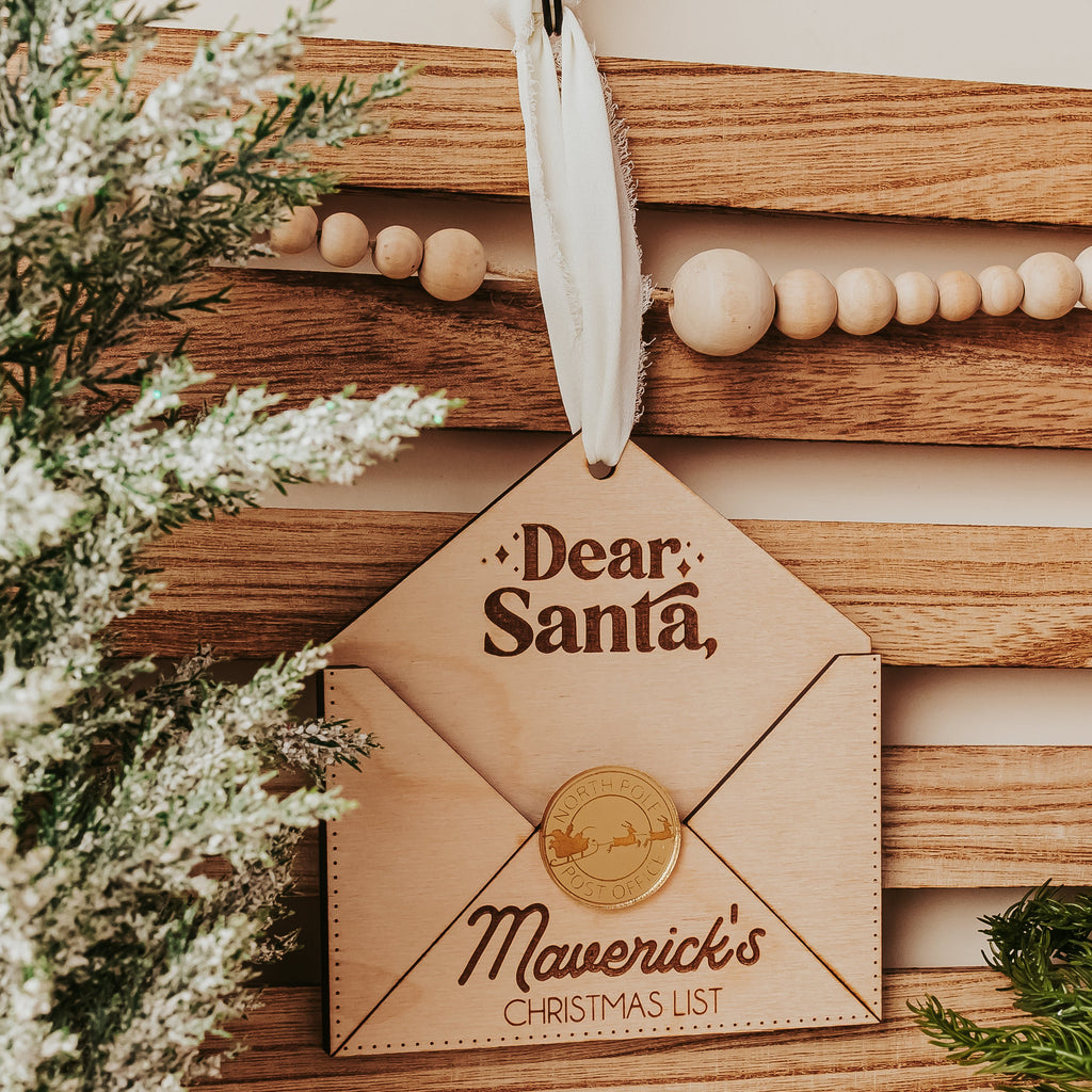 A wooden Christmas envelopeornament designed to look like a letter envelope, hanging from a white ribbon. The ornament features the text 'Dear, Santa,' and 'Maverick's CHRISTMAS LIST' with a printed gold seal.