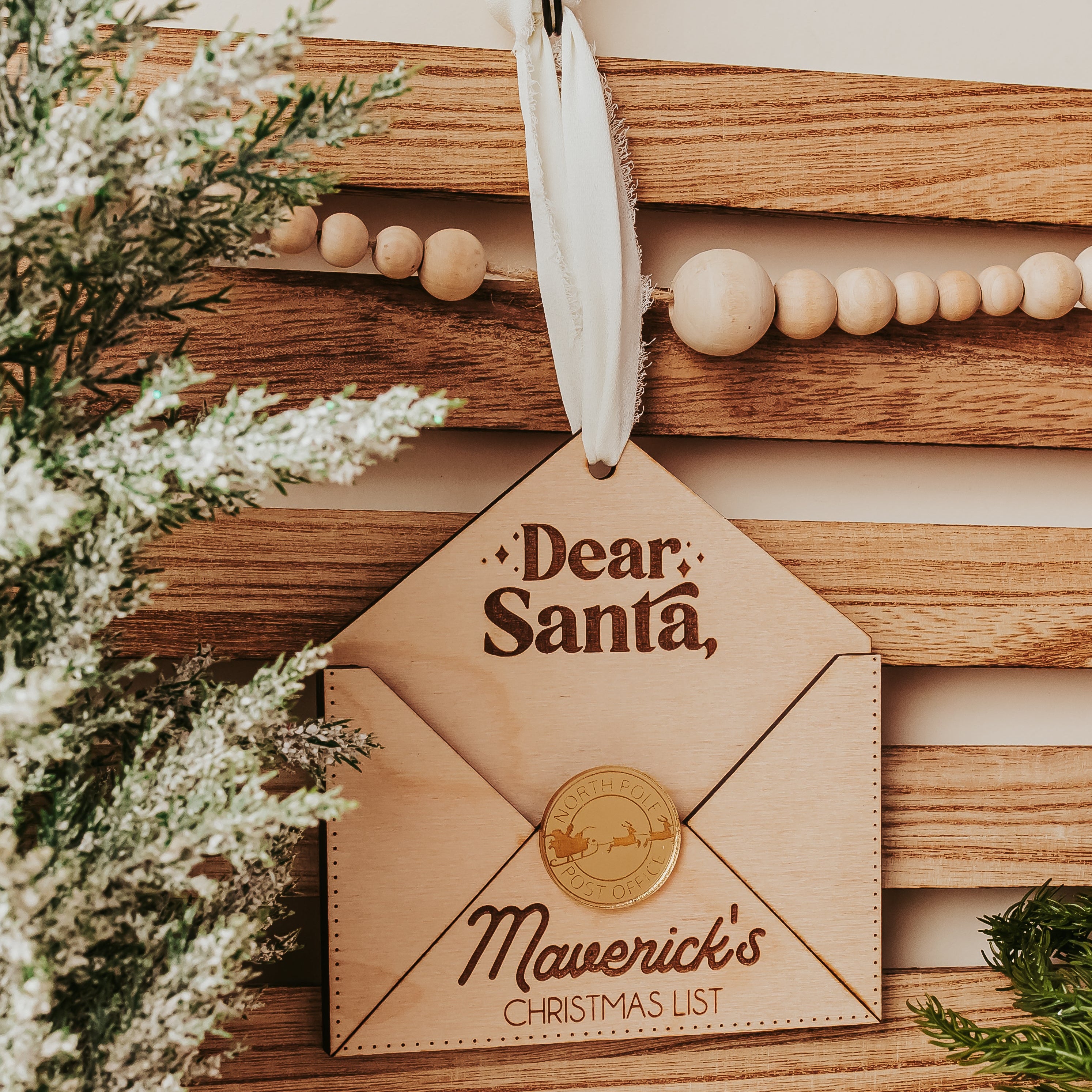 A wooden Christmas envelopeornament designed to look like a letter envelope, hanging from a white ribbon. The ornament features the text 'Dear, Santa,' and 'Maverick's CHRISTMAS LIST' with a printed gold seal.
