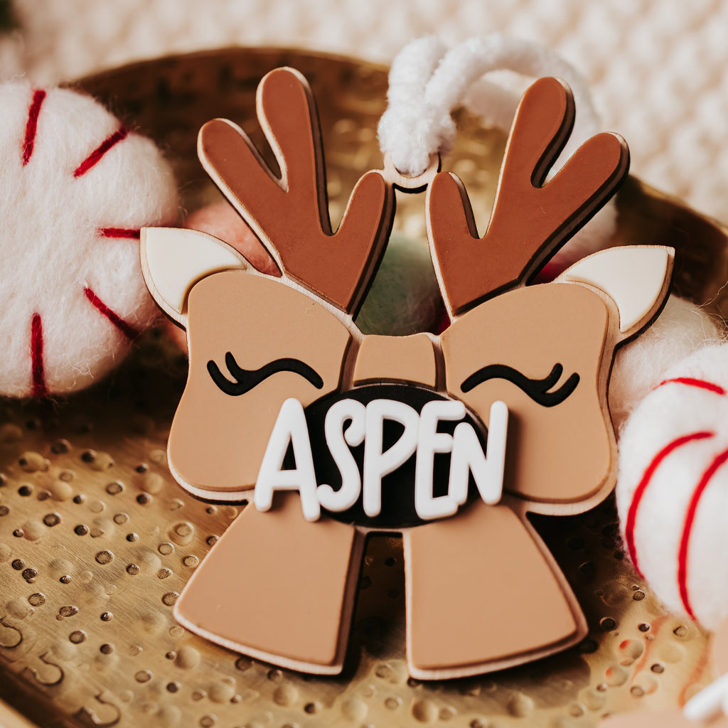 A close-up of a Christmas ornament shaped like a reindeer themed bow with antlers and the name 'ASPEN' in white letters on a black background. The ornament is styled on a metallic tray with holiday-themed props.