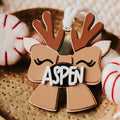 A close-up of a Christmas ornament shaped like a reindeer themed bow with antlers and the name 'ASPEN' in white letters on a black background. The ornament is styled on a metallic tray with holiday-themed props.
