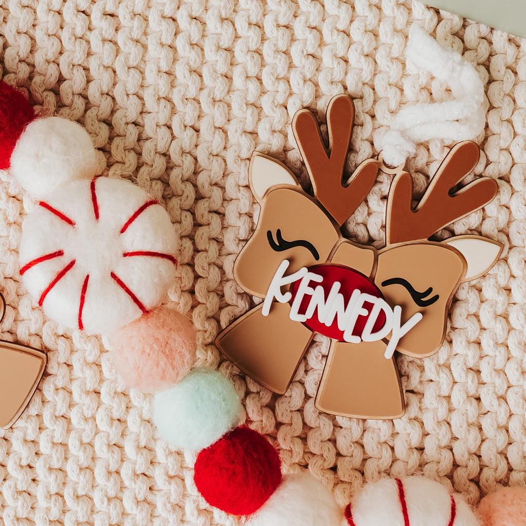 Personalized Christmas ornament featuring two wood and acrylic reindeer themed bow with the name 'KENNEDY' on their chests, decorated with red, white, pink, and mint green felt accents, displayed on a cream-colored knit blanket.