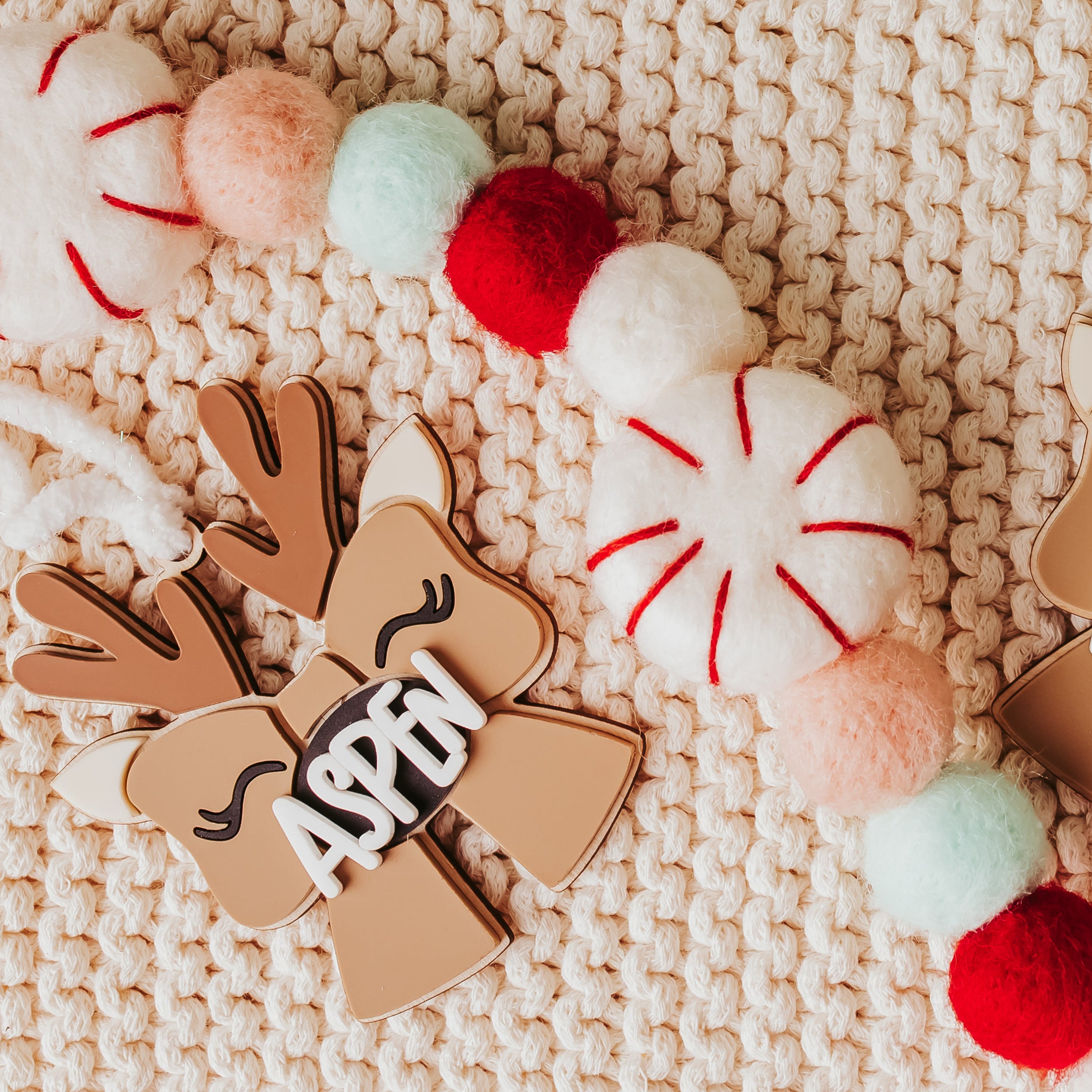 Personalized Christmas garland featuring wood and acrylic reindeer  themed bow ornaments with the names 'ASPEN' their chests, decorated with red, white, pink, and mint green felt accents, displayed on a cream-colored knit blanket.