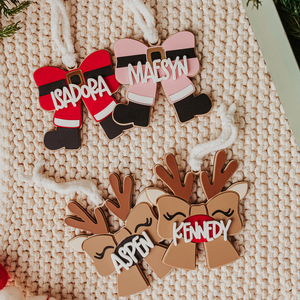 Personalized Christmas ornaments displayed on a beige knit blanket, featuring two Santa-themed bow ornaments with names 'ISADORA' and 'MAESYN' in red and pink, and two reindeer-themed bow ornaments with names 'ASPEN' and 'KENNEDY' in brown. Decorative green pine branches and a small decorated tree are visible around the ornaments.