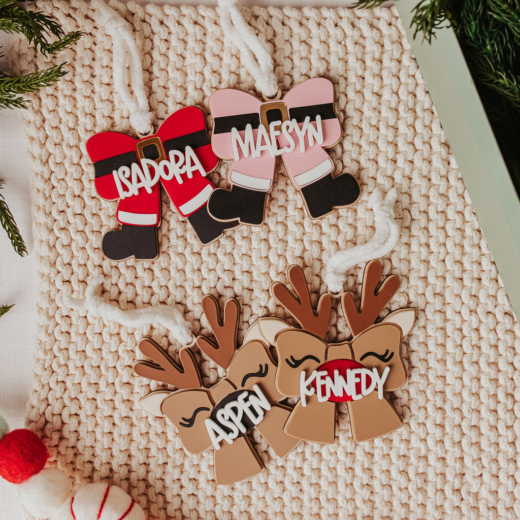 Personalized Christmas ornaments displayed on a beige knit blanket, featuring two Santa-themed ornaments with names 'ISADORA' and 'MAESYN' in red and pink, and two reindeer-themed ornaments with names 'ASPEN' and 'KENNEDY' in brown. Decorative green pine branches and a small decorated tree are visible around the ornaments.