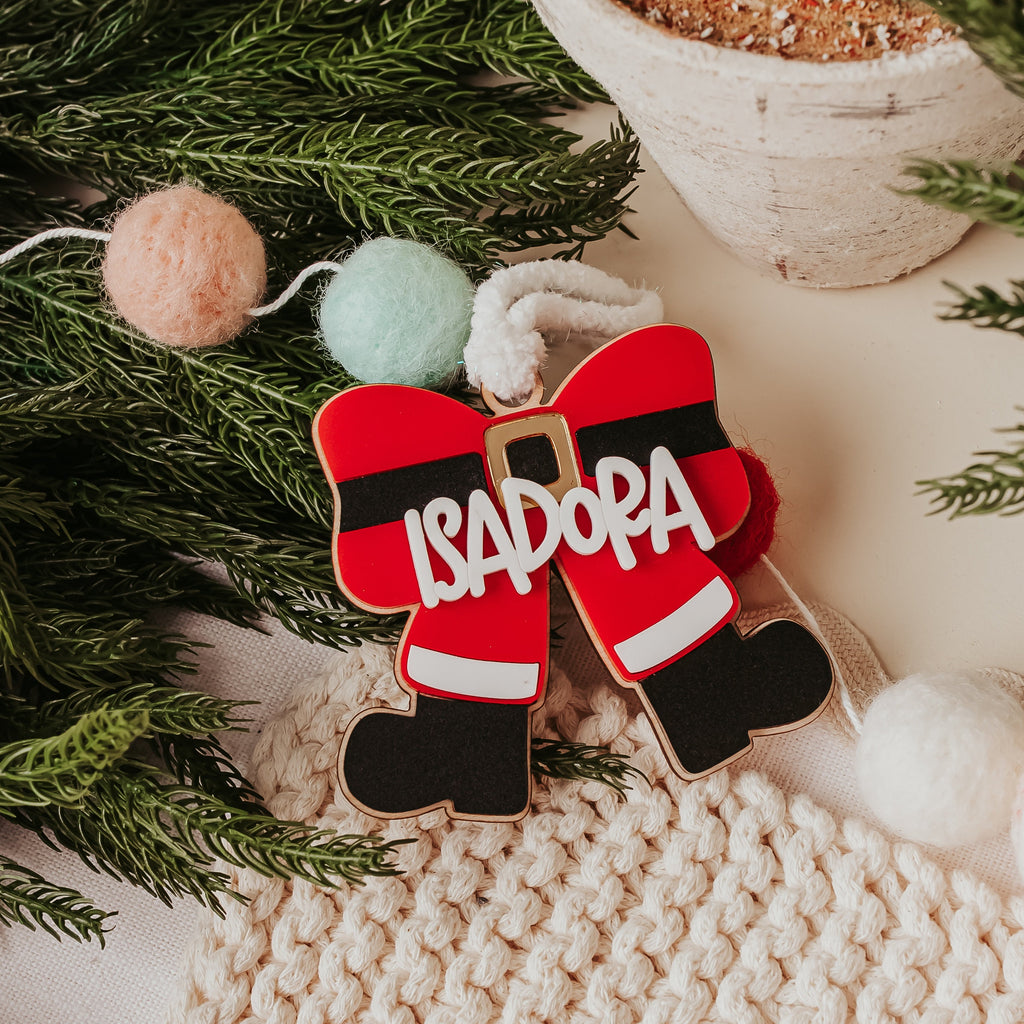 Personalized Christmas ornament bow shaped like Santa's boots in red, black, and white, with the name 'ISADORA' written in white across the front, styled on green pine branches with pom-poms and a knit blanket.