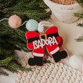 Personalized Christmas ornament bow shaped like Santa's boots in red, black, and white, with the name 'ISADORA' written in white across the front, styled on green pine branches with pom-poms and a knit blanket.