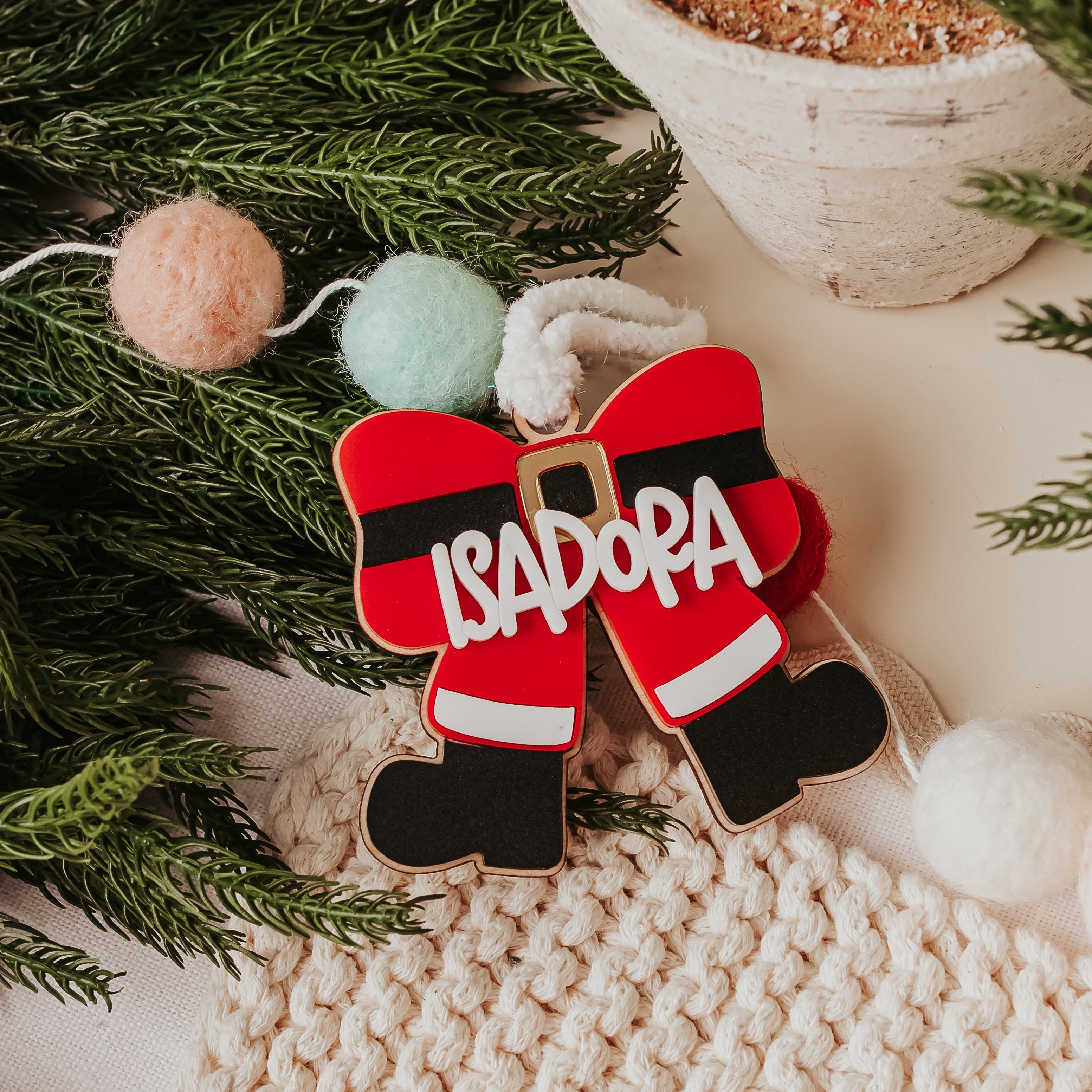 Personalized Christmas ornament bow shaped like Santa's boots in red, black, and white, with the name 'ISADORA' written in white across the front, styled on green pine branches with pom-poms and a knit blanket.