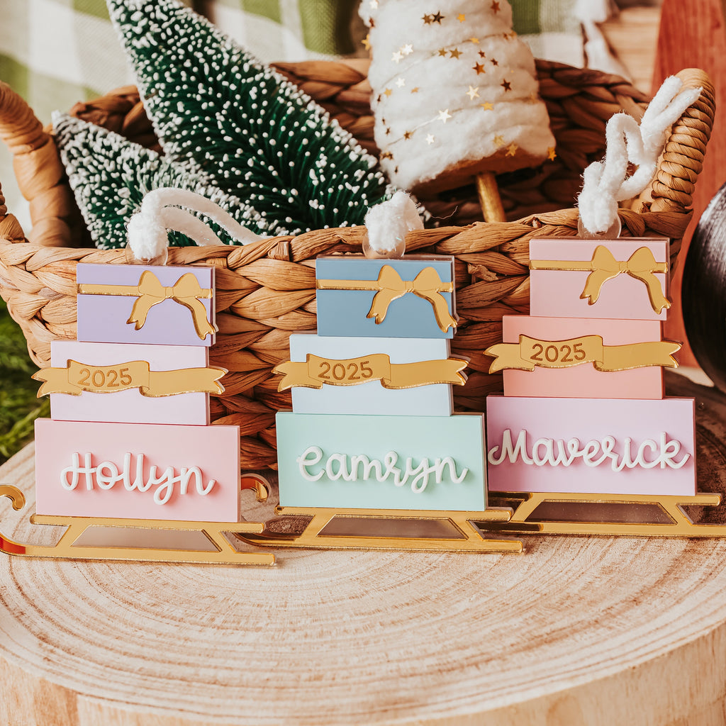 Three versions of a gold sled ornament stacked with pastel gift box with gold ribbon accents, displayed on a rustic wood slice with holiday decorations in the background.